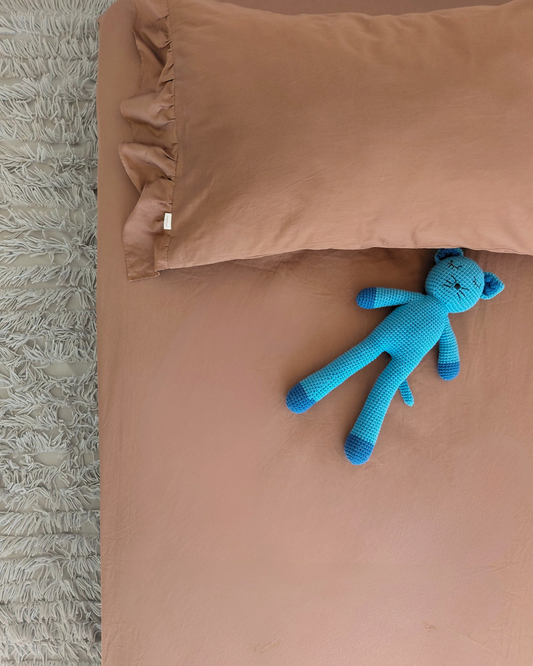 A close-up of a bed with a solid Small Batch Bedding cotton chocolate brown fitted sheet and matching pillowcase with one-sided ruffle, styled with a small blue plush toy on the sheet and a textured gray rug on the side.