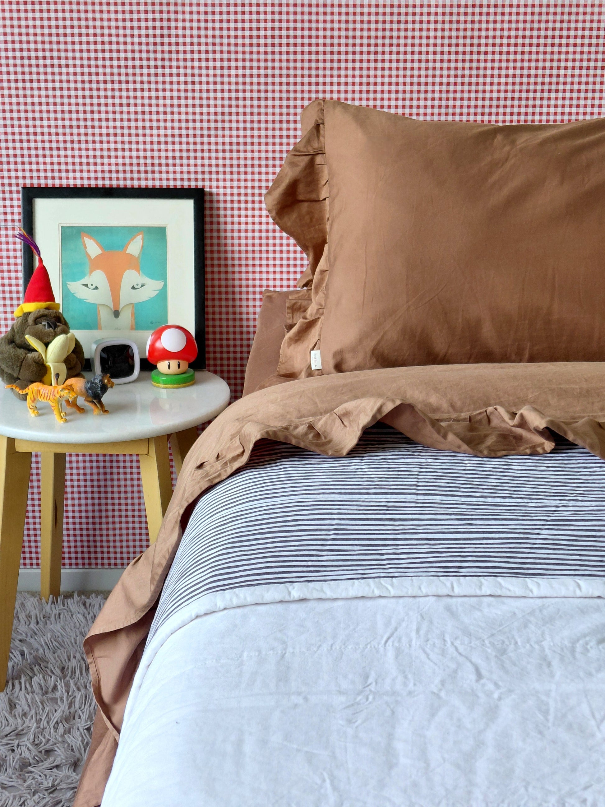 A styled bedroom scene showing a bed with Small Batch Bedding brown ruffled cotton pillowcases and matching brown ruffle cotton sheet, layered over a black-and-white striped sheet and a light gray duvet. A small side table with toys and a framed fox print sits beside the bed against a pink-and-white checkered wall.