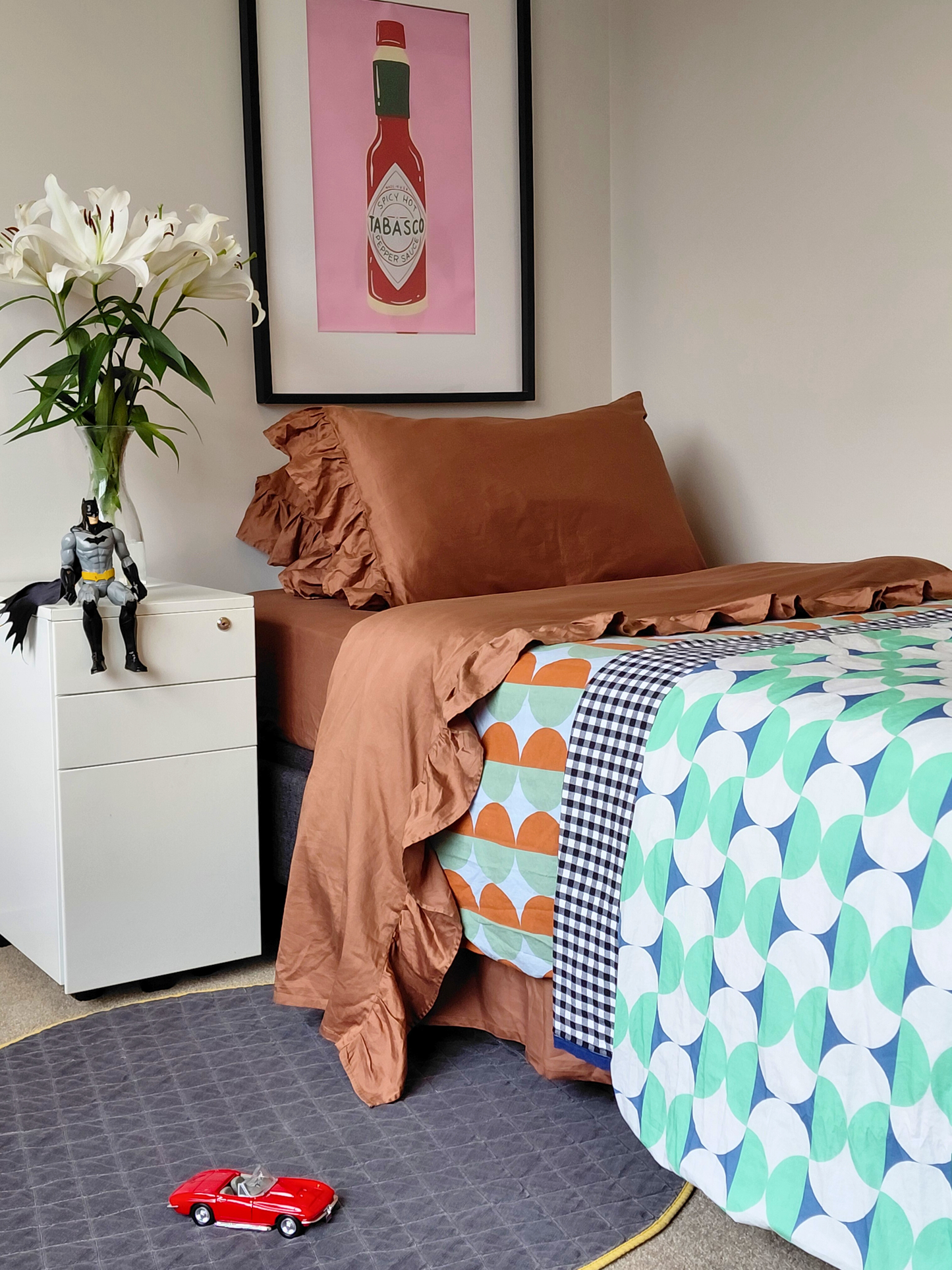 Bedroom with chocolate brown cotton flat sheet and pillowcases with patterned bedspread, white nightstand, and colorful decor.