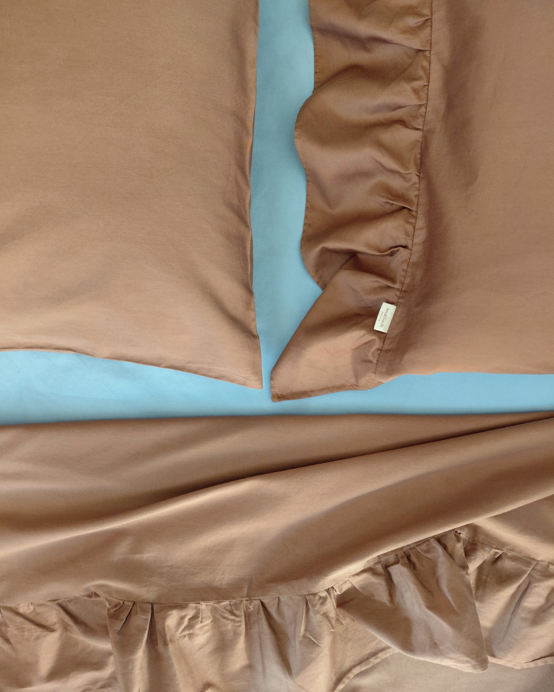 Chocolate brown cotton pillowcases with one sided ruffle and chocolate brown cotton top sheet with ruffle on a light blue fitted sheet, showing a brand label for Small Batch Bedding.