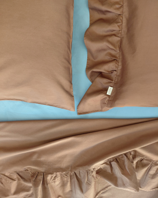 Chocolate brown cotton pillowcases with one sided ruffle and chocolate brown cotton top sheet with ruffle on a light blue fitted sheet, showing a brand label for Small Batch Bedding.