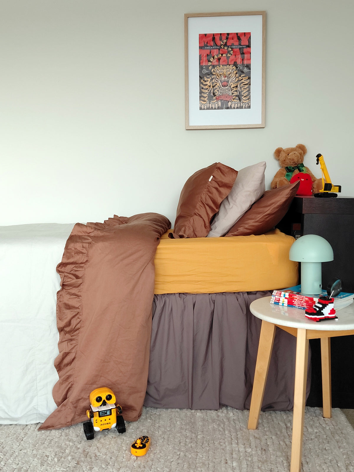 A styled bedroom corner showing a bed with brown ruffled bedding, a mustard yellow center panel, a gray skirted bed skirt, a small white side table, and toys on a carpeted floor.
