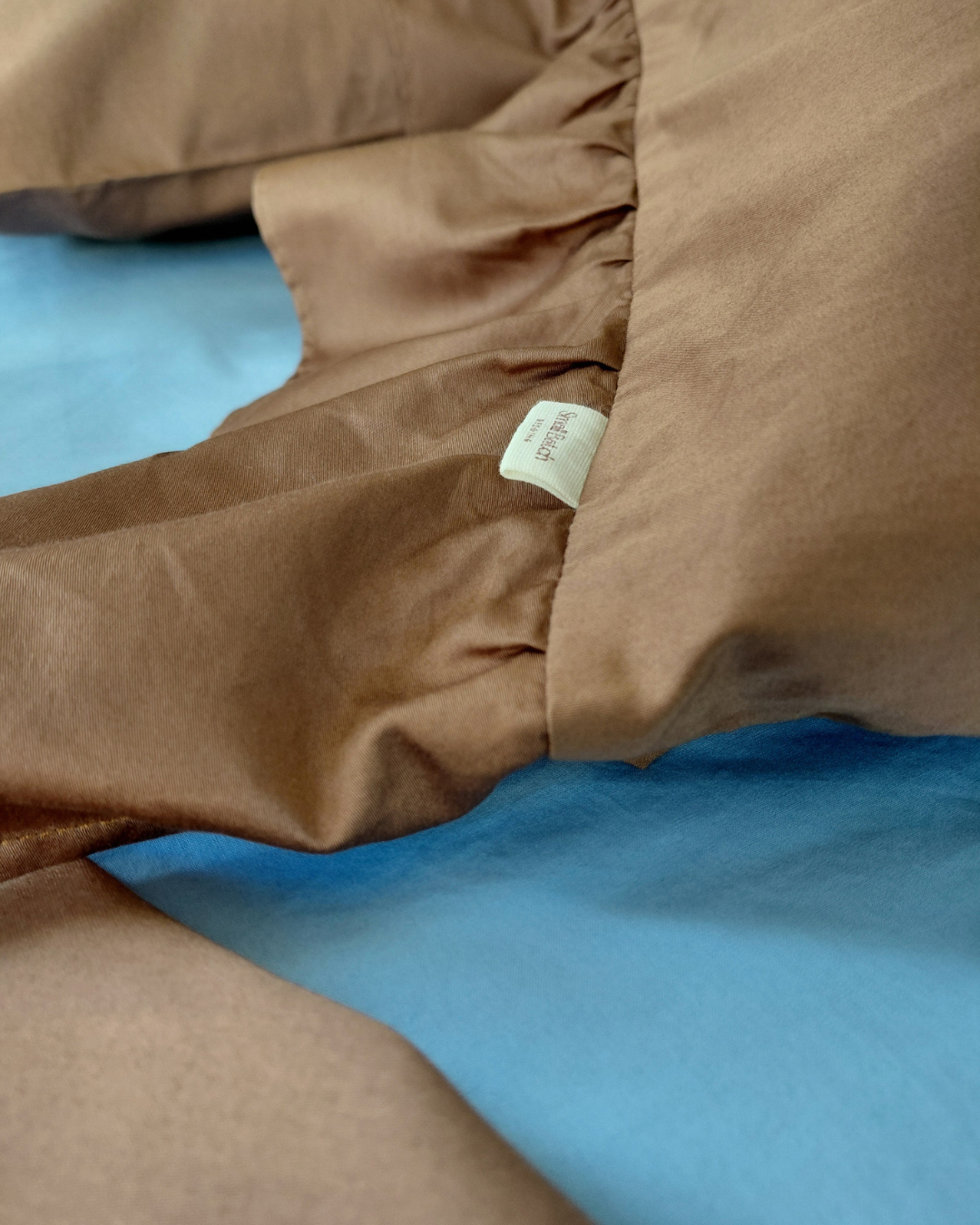 Brown cotton pillowcase ruffle with a Small Batch Bedding brand tag on a blue background