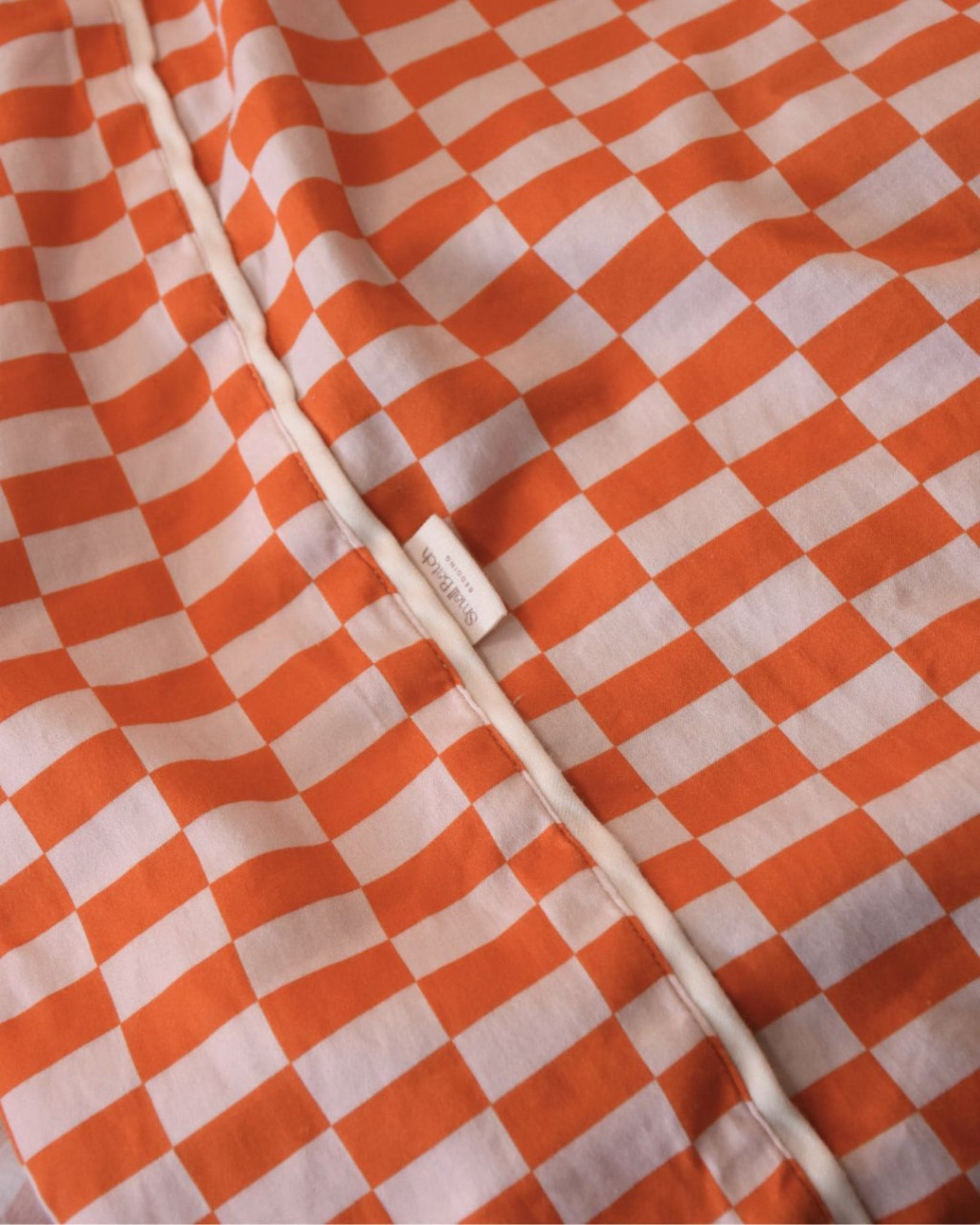 Close-up of tomato red orange and light rectangle checkered cotton fitted sheet fabric with Small Batch Bedding brand label. Kid's bedding single sheet.
