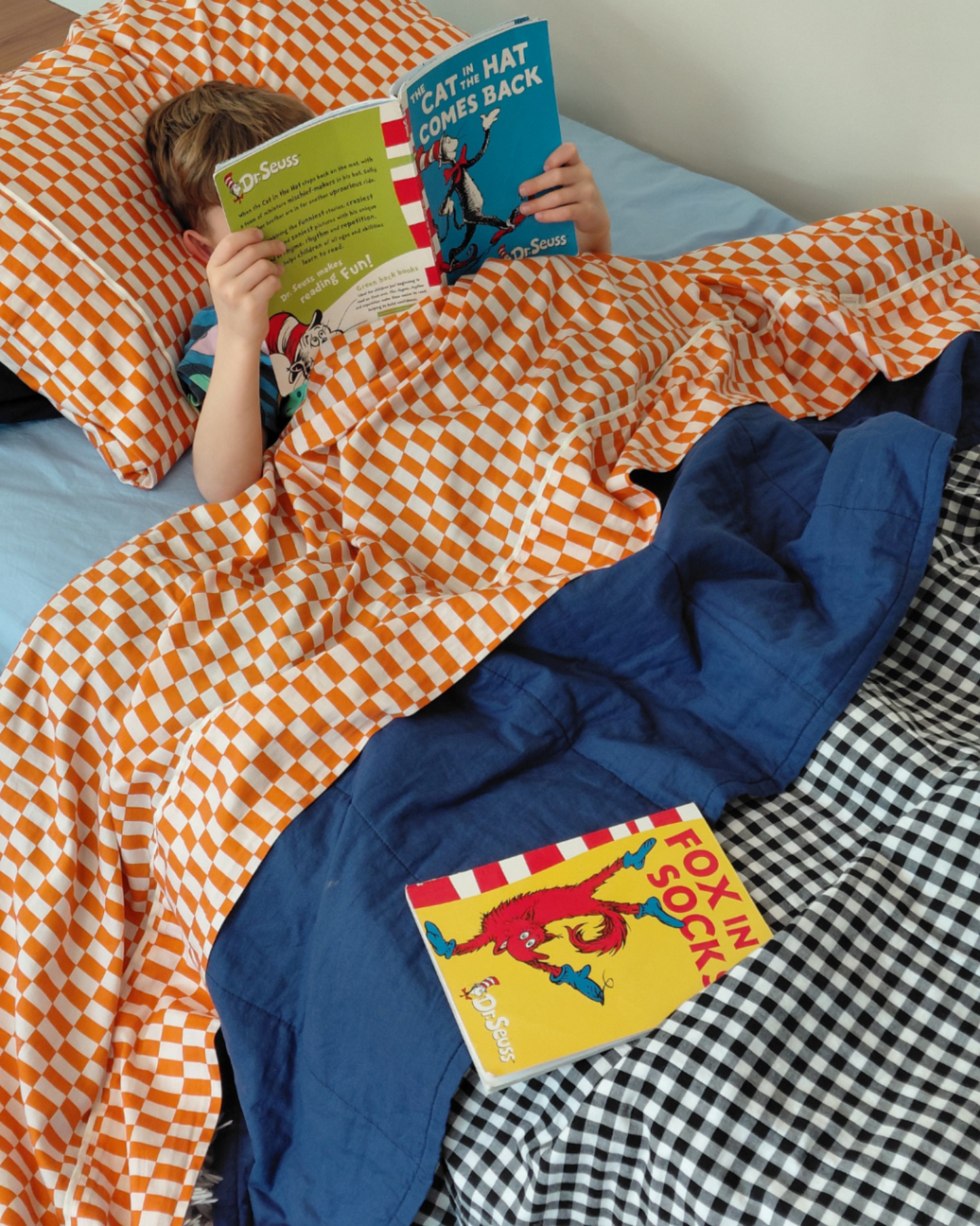 Child reading a book under orange checkered tomato tile cotton sheets and a cobalt blue cotton quilt with gingham blank and white reverse with another book on the bed.