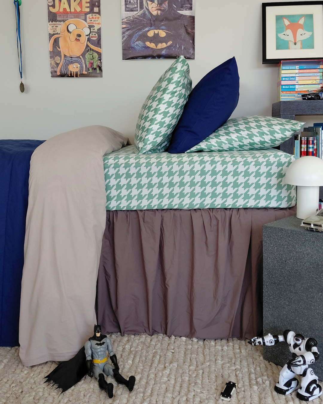 Bedroom with a bed featuring green and white houndstooth cotton sheeting, surrounded by various items including a lamp, posters and toys.
