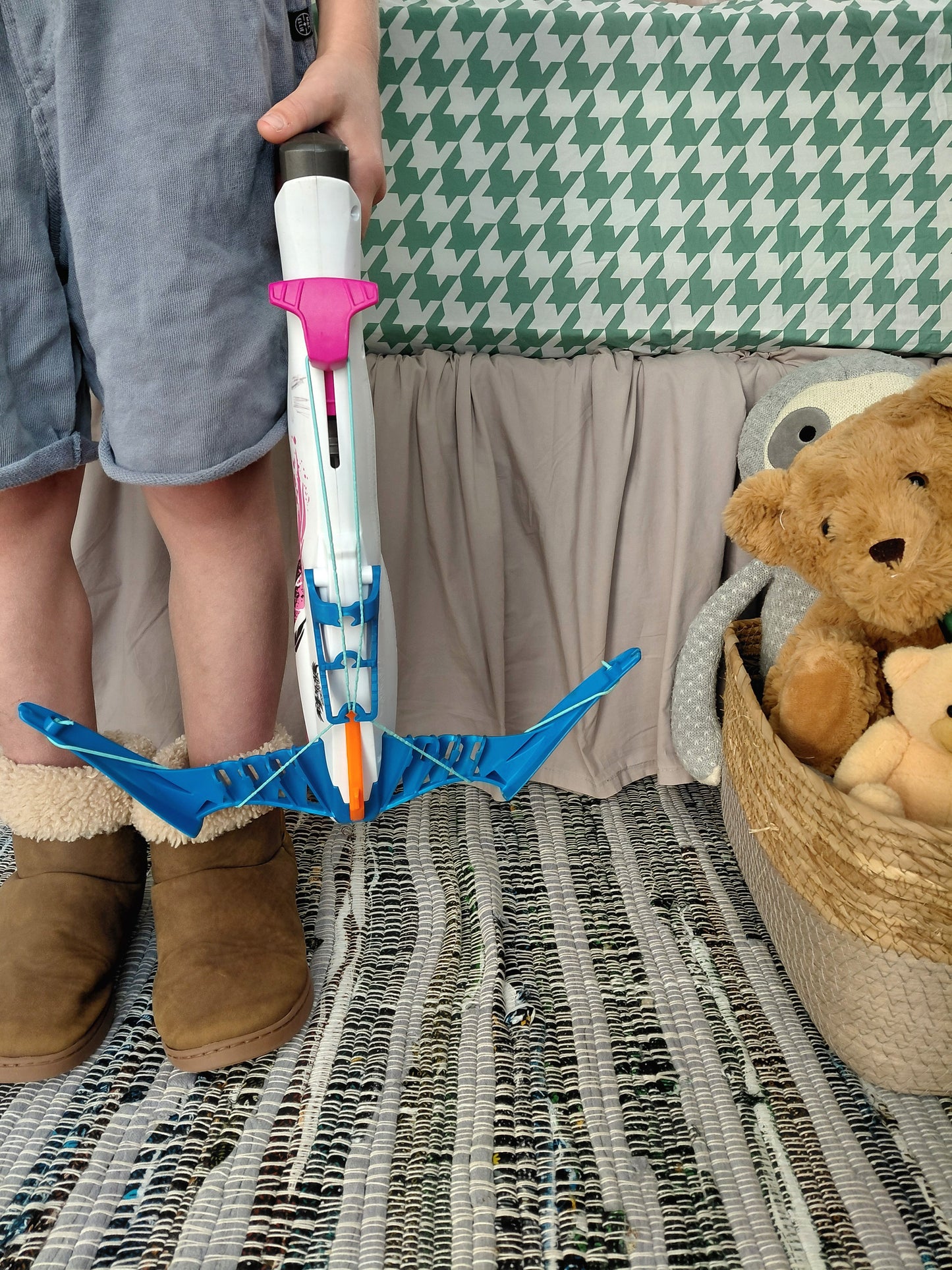 A child holding a toy crossbow with a blue bow and white, blue, and pink arrow, standing on a patterned rug with a teddy bear in a basket in the background in front of a green and white houndstooth fitted sheet.