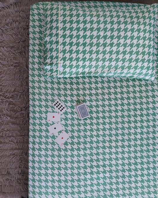 A Small Batch Bedding 100% cotton green and white houndstooth patterned fitted sheet set with a matching pillowcase, laid flat on a bed alongside four playing cards. The bed is positioned next to a brown shag rug.