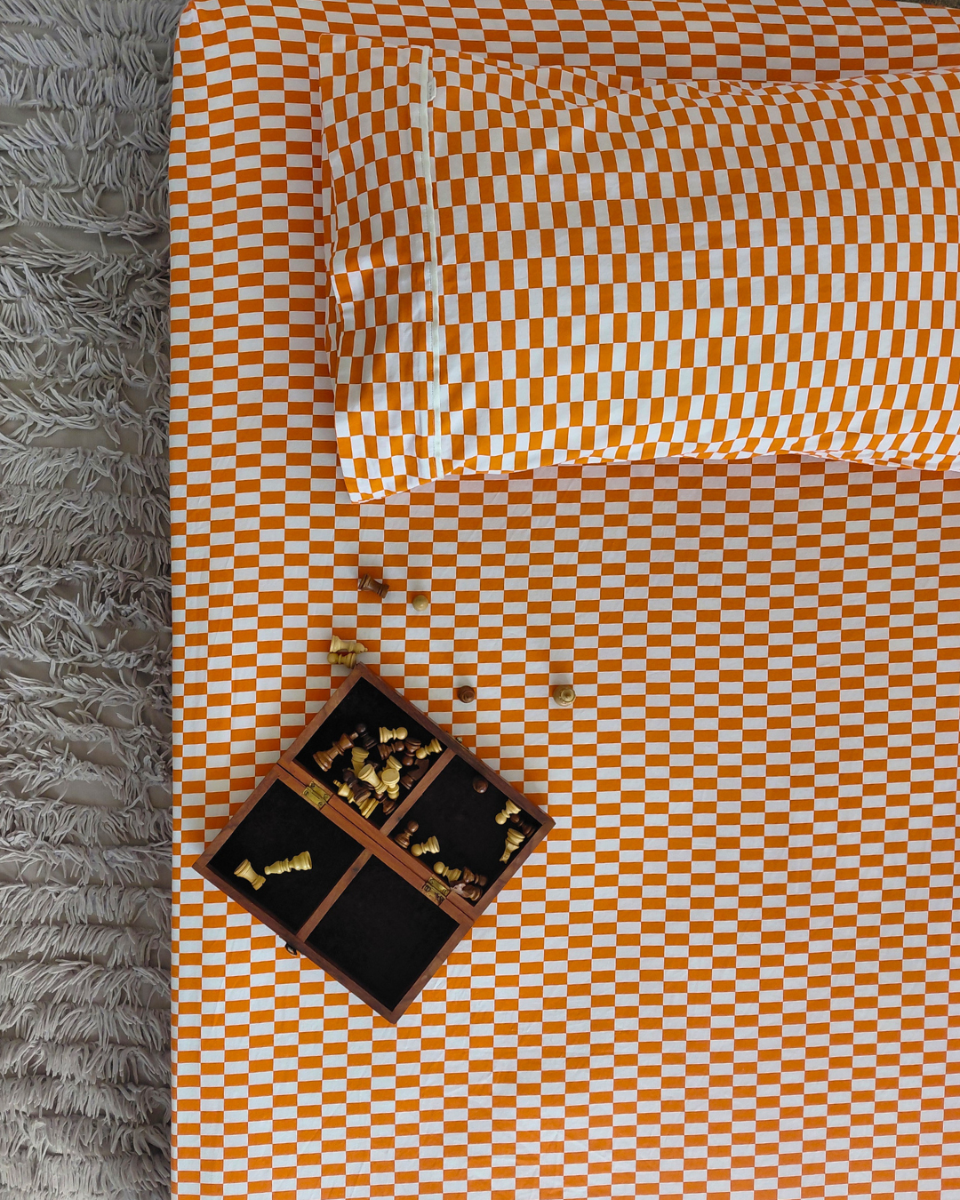 An overhead view of a bed dressed in a Small Batch Bedding red tomato tile cotton patterned fitted sheet and pillowcase, styled with a small wooden box of checkers on a gray shag rug.