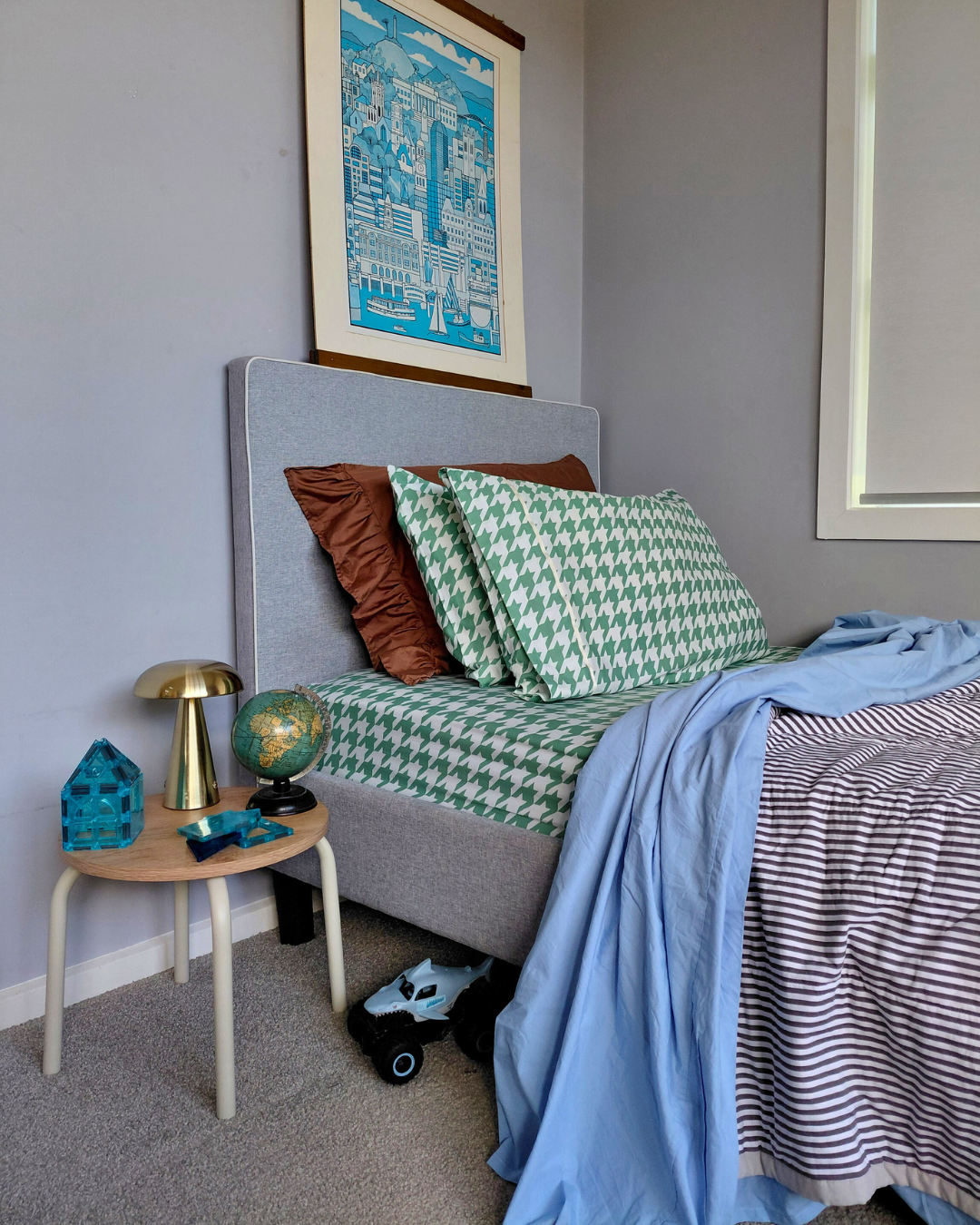 Bedroom with a grey bed featuring green and white checkered houndstooth cotton bedding, brown ruffle and houndstooth green patterned pillows, and a stiped black and white blanket.