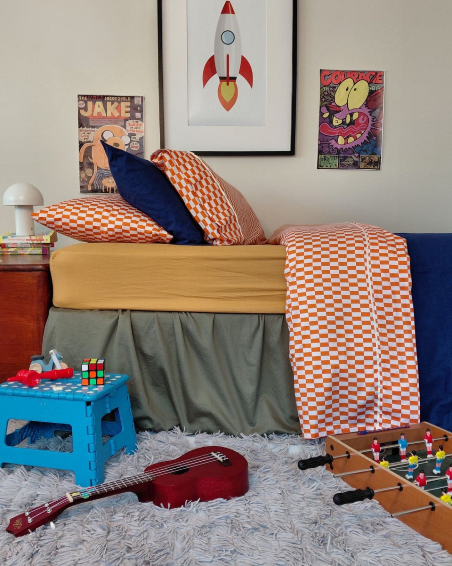 Children's bedroom styled with made bed, toys, and wall art, tomato red orange cotton flat sheet, matching pillowcases with royal blue blanket on a gold fitted sheet and green valance.  kids sheets.