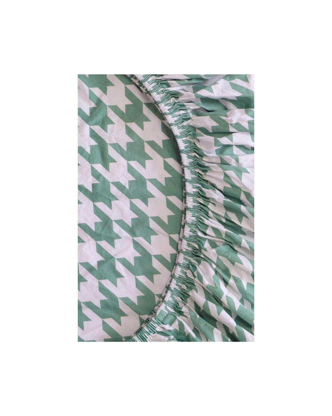 A close-up of a green and white houndstooth patterned fitted sheet with a gathered elasticized edge, shown against a plain white background.