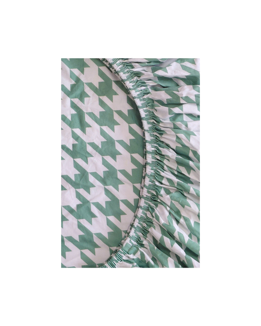 A close-up of a green and white houndstooth patterned fitted sheet with a gathered elasticized edge, shown against a plain white background.