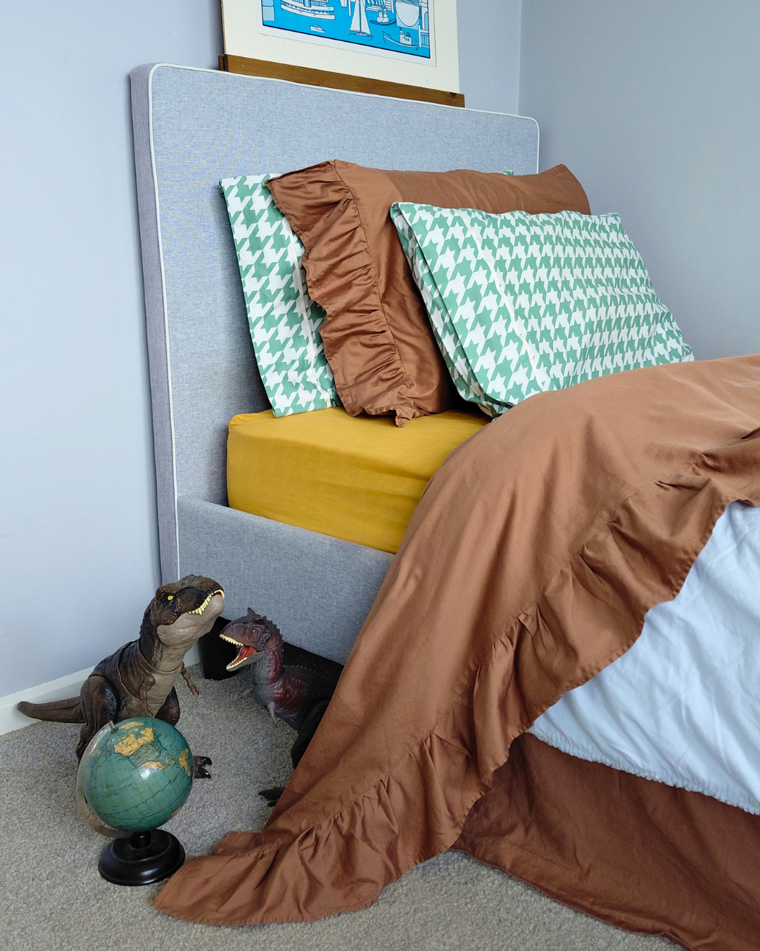 Bed with brown ruffle top sheet and colorful pillows including green and white houndstooth pillows, next to a globe and toy dinosaurs on a gray carpet.