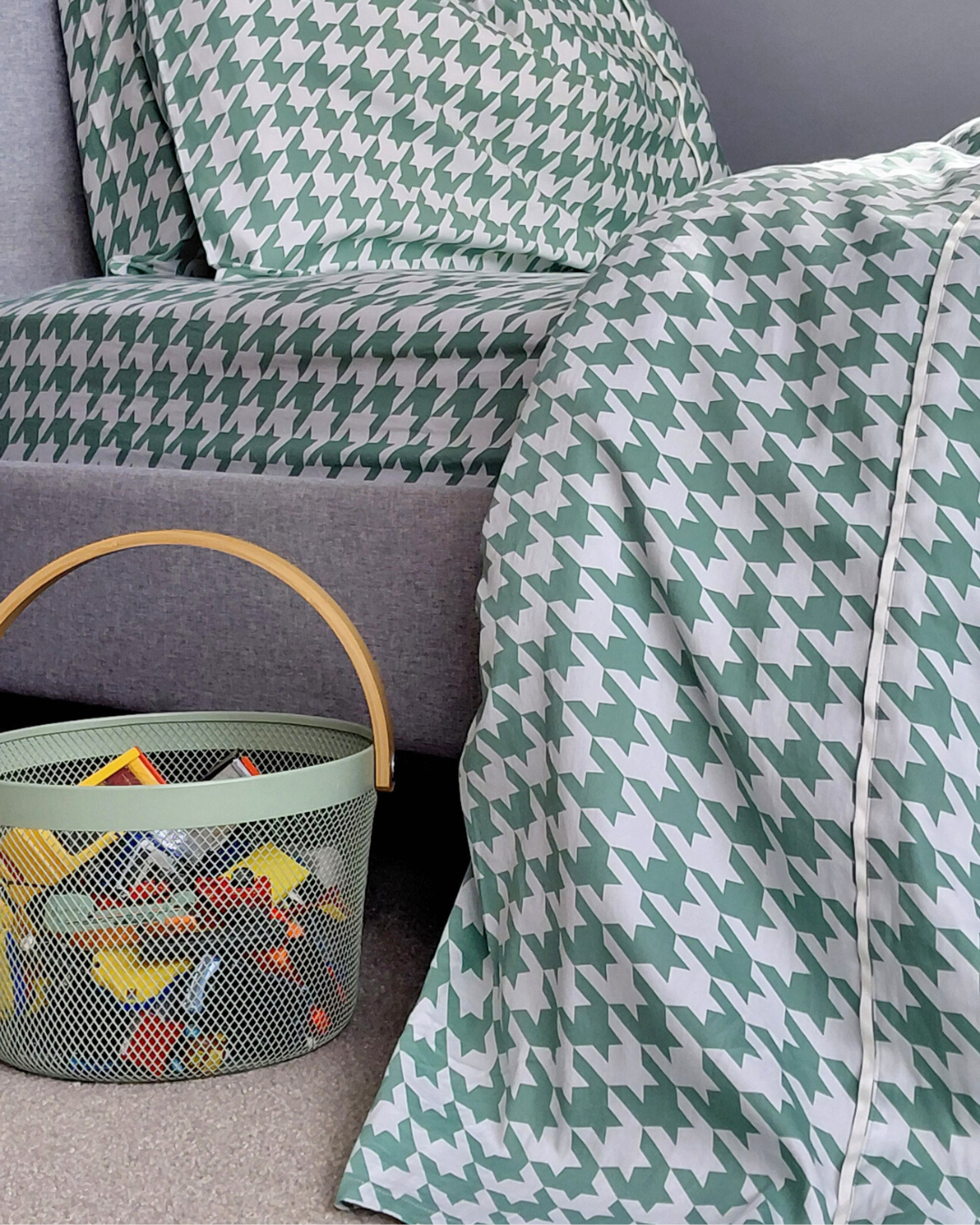 Pea green and white houndstooth patterned cotton sheet draped over a bed with a basket in the foreground.