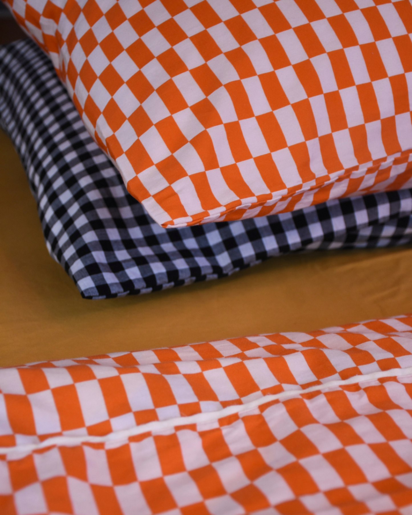 Close up of tomato red and orange checkered tile cotton pillowcase with matching flat red orange checkered sheet by Small Batch Bedding, Styled with gold flat sheet and black gingham pillowcase.