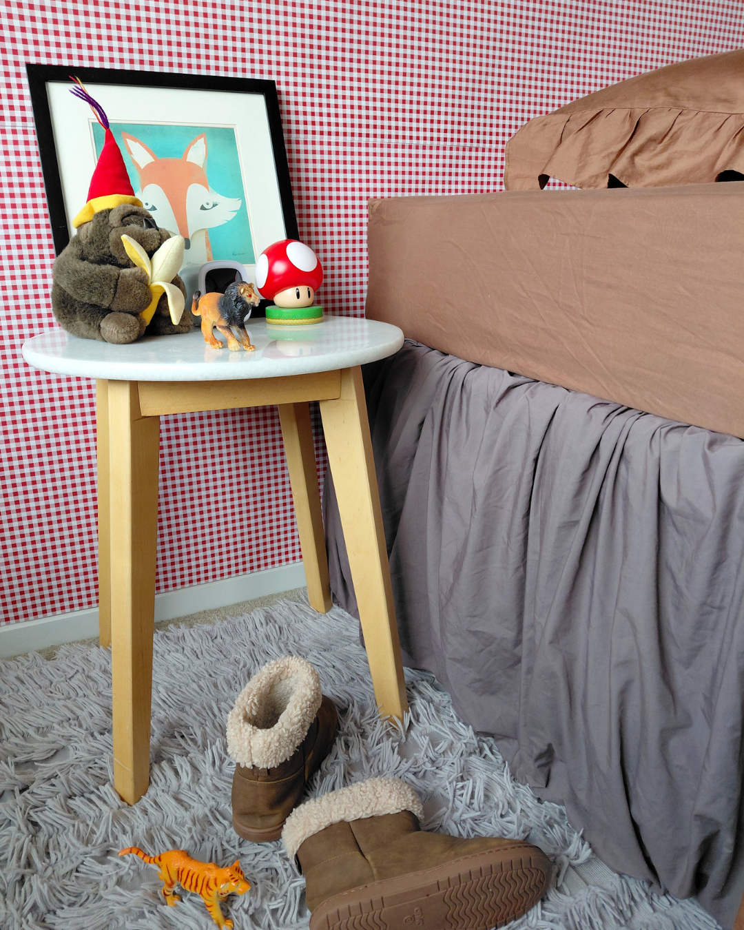 A styled bedroom corner showing a small round white side table with light wood legs, decorated with plush toys and a framed picture, next to a bed with a Small Batch Bedding chocolate brown fitted sheet and one-sided chocolate ruffle pillowcase. The bed has a gray skirt. A pair of brown shearling-lined boots and a small toy are on a gray shag rug against a red-and-white checkered wall.