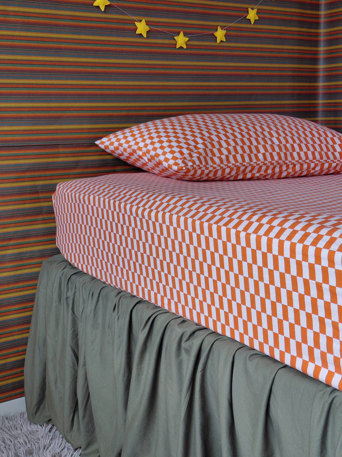 A bed dressed in a fitted red and white tomato tile cotton sheet and pillowcase by Small Batch Bedding, with a dark olive green bed skirt, set against a colorful striped wall with yellow star garlands.