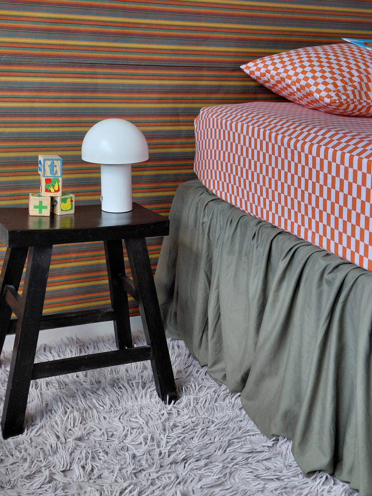 A styled bedroom corner featuring a bed with red and white tomato tile cotton bedding from Small Batch Bedding and a forest green skirt, next to a black wooden stool holding a white mushroom-shaped lamp and colorful blocks, set against a colorful striped wallpaper.