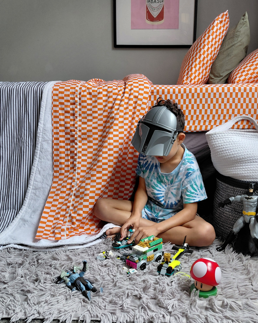 Child playing with toys in a room with a red tomato tile checkered top and bottom cotton sheet, white and black and white striped cotton blanket and framed picture on the wall.