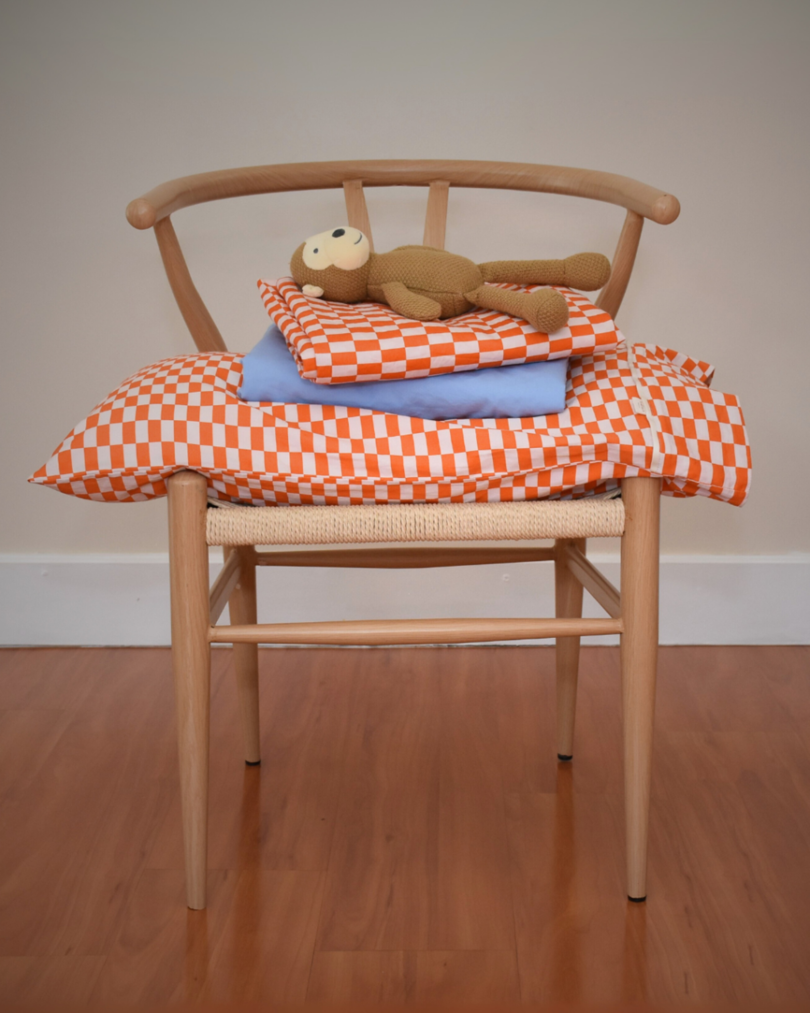 Wishbone chair with tomato red orange checkered cotton pillowcase, blue sheet and tomato red orange checkered flat sheet with a teddy bear on a plain background and wooden floor.