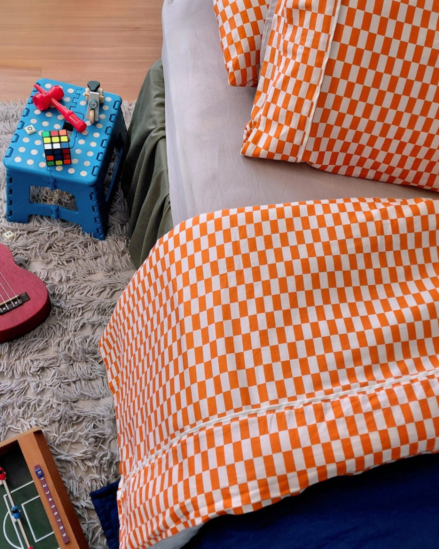 Red orange and white checkered fitted cotton sheet with matching tomato tile pillowcases on a child's bed with toys and a small table in the background