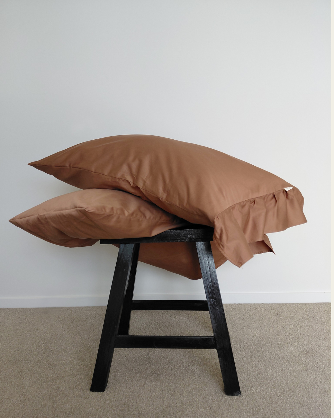 Brown cotton pillows with ruffle on a black stool against a white wall.