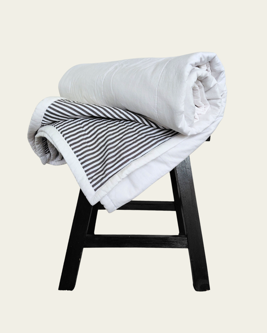 Folded white cotton blanket with striped patterned inner on a wooden stool against a beige background